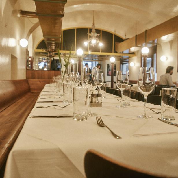 Elegant gedeckter Restauranttisch mit Gläsern und Besteck in warm beleuchtetem Speisesaal, im Hintergrund sitzen zwei Gäste.