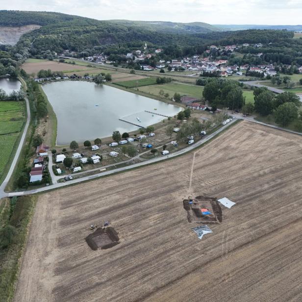 Luftaufnahme eines Dorfes mit See, Feldern, Hügeln und einer archäologischen Ausgrabung im Vordergrund.