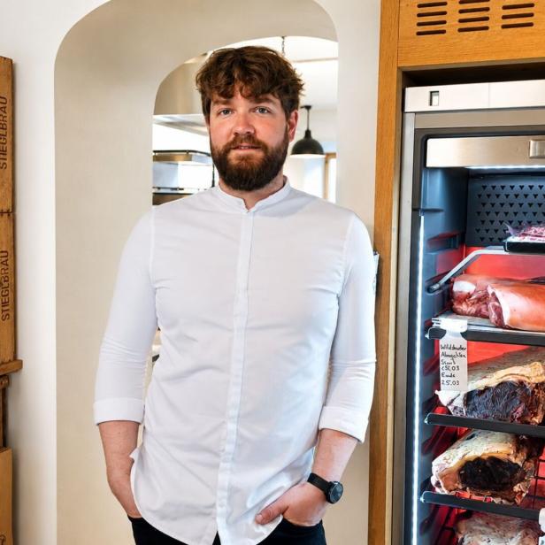 Ein bärtiger Mann in einem weißen Hemd steht vor einem Dry-Ager-Kühlschrank mit Fleischstücken neben einem Regal mit Stieglbräu-Flaschen.