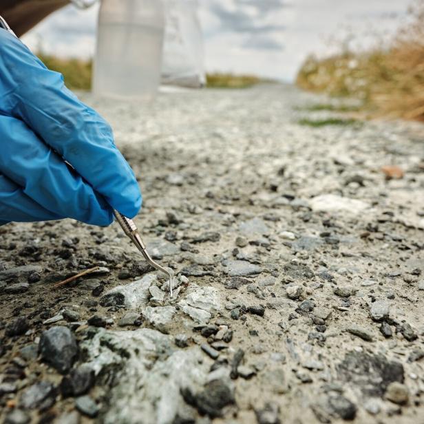 Behandschuhte Hand untersucht mit einem Metallwerkzeug den rissigen Asphalt am Straßenrand.