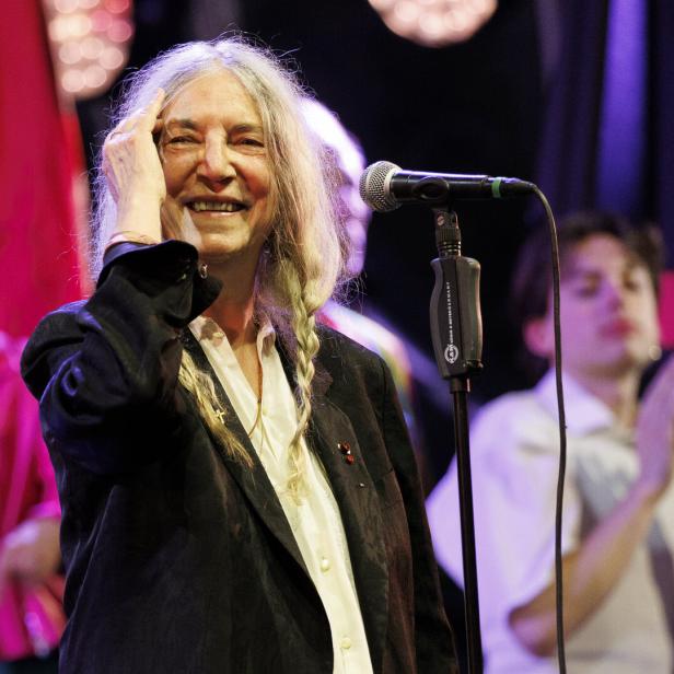 Patti Smith lächelt auf einer Bühne vor einem Mikrofon, während sie sich mit der Hand durch ihr langes graues Haar fährt.