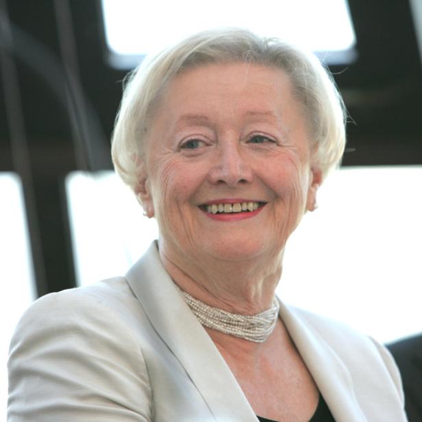 Lächelnde Karin Kathrein mit weißem Haar in heller Jacke und Perlenkette vor hellem Hintergrund.