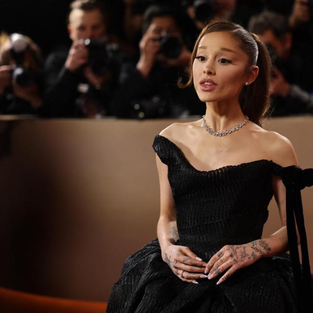 Ariana Grande trägt ein schwarzes schulterfreies Kleid und eine Halskette vor einer Kulisse aus Fotografen.