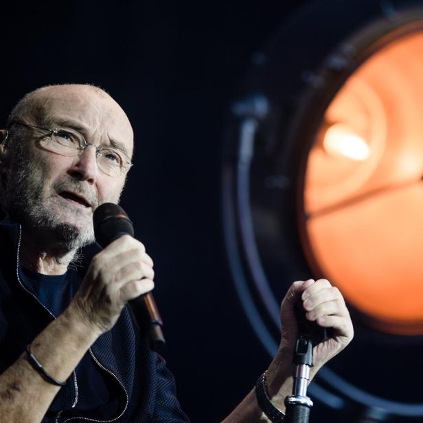 Phil Collins singt vor einem großen, orange leuchtenden Scheinwerfer in ein Mikrofon.
