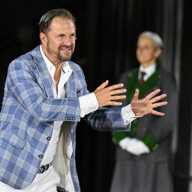 Philipp Hochmair in einem blau karierten Sakko gestikuliert vor einer Person in traditioneller Tracht.