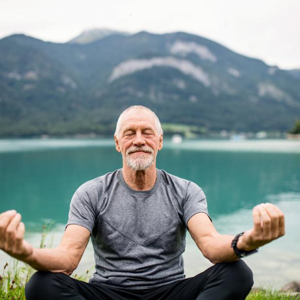 A senior man pensioner sitting by lake in nature, doing yoga exercise.