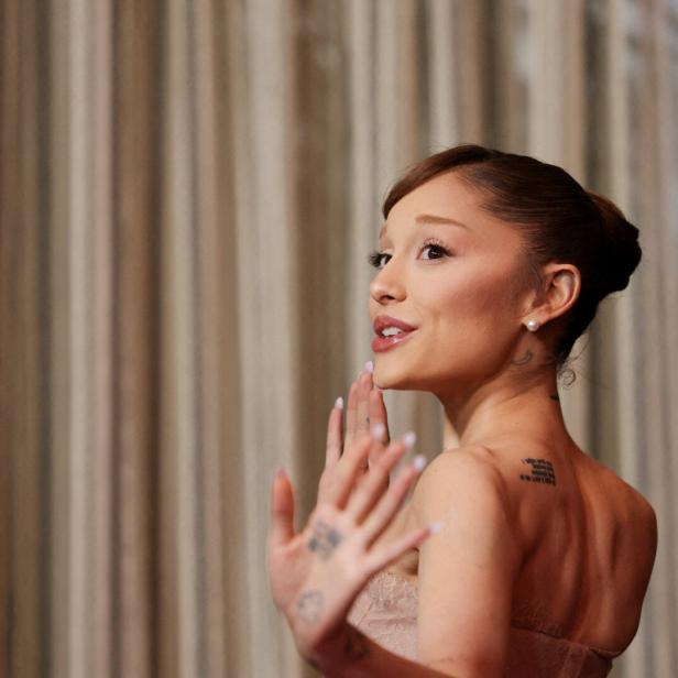 Ariana Grande blickt über die Schulter und hebt die Hand vor einem hellen Vorhang.