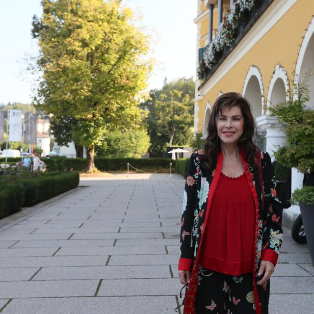 Eine Frau im roten Oberteil und floralen Mantel steht vor einem gelben Gebäude mit Bögen.