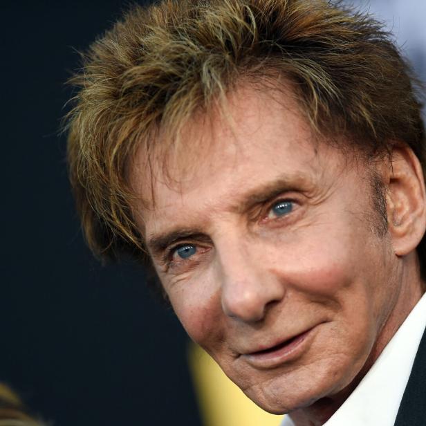 Nahaufnahme des Sängers Barry Manilow mit seinem charakteristischen blonden Haar vor einem dunklen Hintergrund.