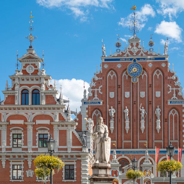 Das Schwarzhäupterhaus in Riga mit seiner verzierten Backsteinfassade und einer davorstehenden Roland-Statue unter blauem Himmel.