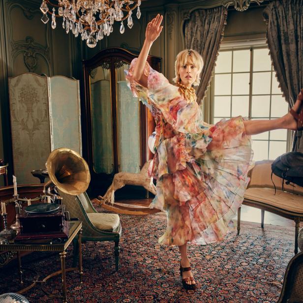 Eine Frau in luftiger Blumenkleidung tanzt mit erhobenem Bein in einem opulent eingerichteten Schlafzimmer.