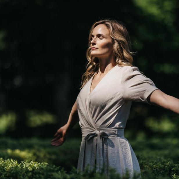 Eine Frau mit blondem Haar steht in einem hellen Kleid mit geschlossenen Augen und ausgebreiteten Armen in einem sonnigen Garten.