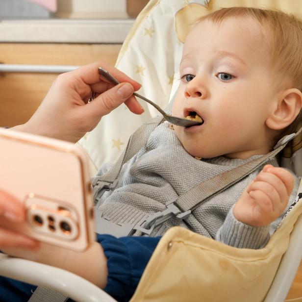 Portrait of cute baby boy eating porridge in highchair and watching cartoons on smartphone