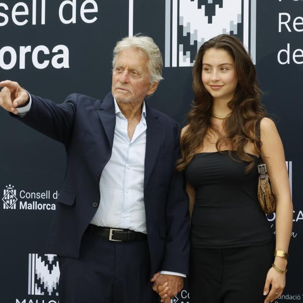 Michael Douglas steht vor einem Banner des Consell de Mallorca und zeigt mit der Hand in die Ferne, während er die Hand Tochter Carys Zeta Jones hält.