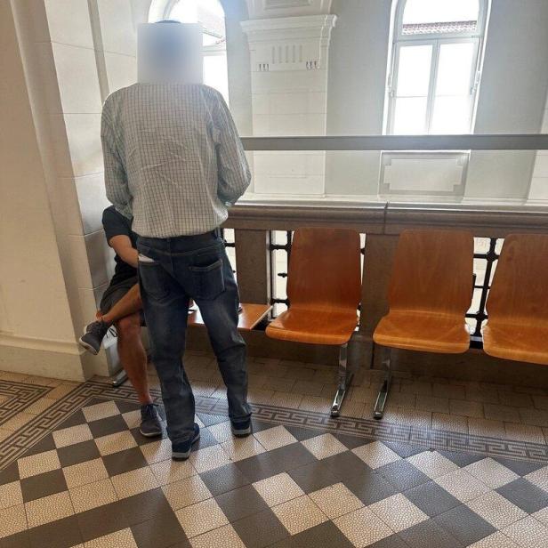 Zwei Männer stehen in einem hellen Flur neben einer Sitzreihe mit Holzstühlen.