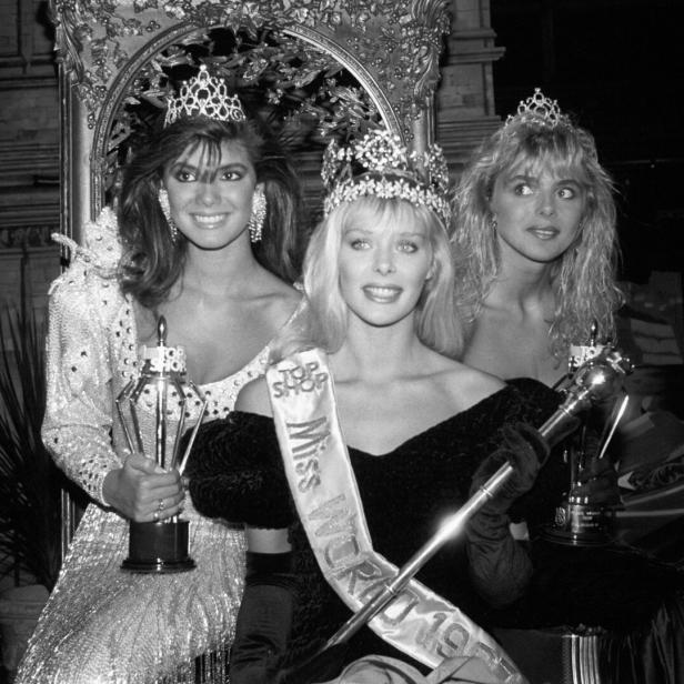 Die Miss World 1987, Ulla Weigerstorfer, sitzt mit zwei weiteren Finalistinnen in Abendkleidern und Kronen auf einer Bühne.