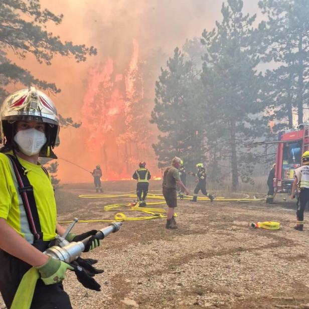 Feuerwehrkräfte bekämpfen einen Waldbrand mit Schläuchen und Ausrüstung, während im Hintergrund Flammen und Rauch aufsteigen.