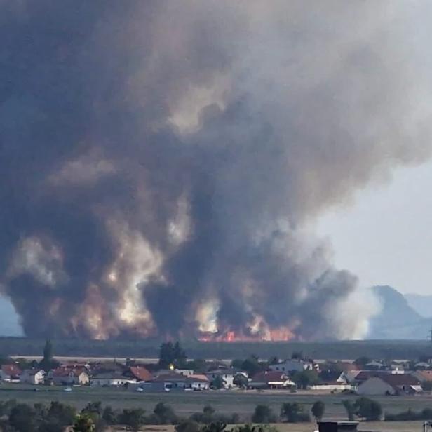 Großbrand mit dichtem schwarzem Rauch über Feldern nahe einer Ortschaft und Bergen im Hintergrund.