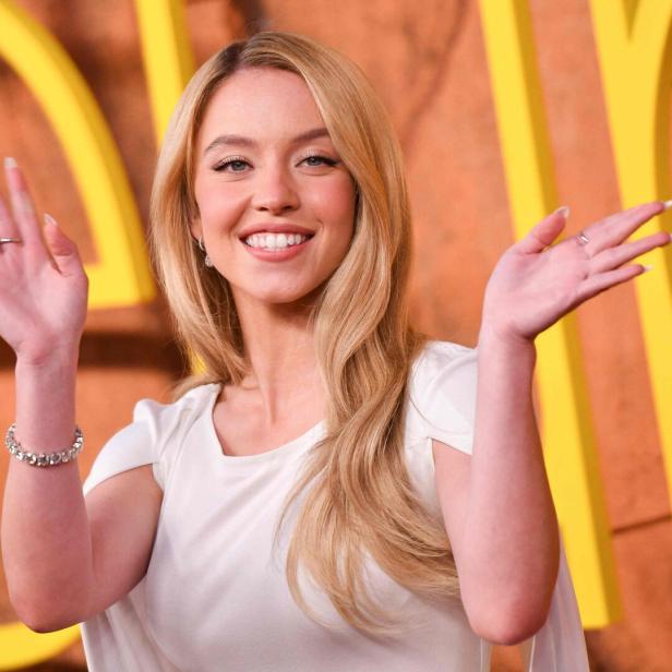 Sydney Sweeney lächelt in die Kamera und winkt mit beiden Händen vor einem gelben Hintergrund.