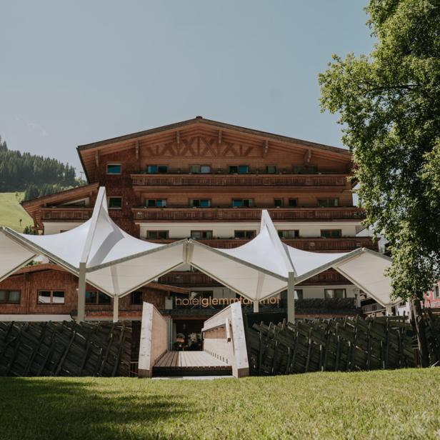 Hotel mit Holzfassade und Sonnensegeln vor grüner Bergkulisse.