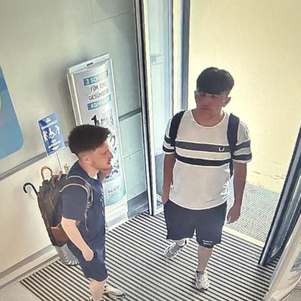 Zwei junge Männer mit Rucksäcken stehen im Eingangsbereich vor einer Glastür.