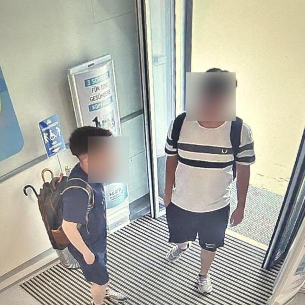 Zwei junge Männer mit Rucksäcken stehen im Eingangsbereich vor einer Glastür.