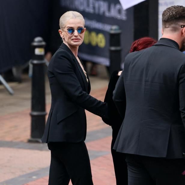 Kelly Osbourne in einem schwarzen Anzug und mit blauer Sonnenbrille geht neben einer Person in einem schwarzen Anzug durch Brindleyplace.