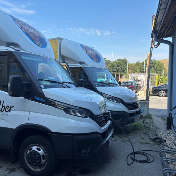 Zwei weiße Elektro-Lieferwagen der Bäckerei Felber stehen an einer blauen Wand und werden an Ladestationen aufgeladen.