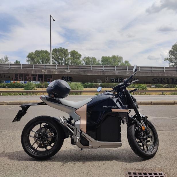 Ein schwarzes Honda-Motorrad mit einem auf dem Sitz liegenden Helm steht an einer Straße vor einer mit Graffiti verzierten Brücke.