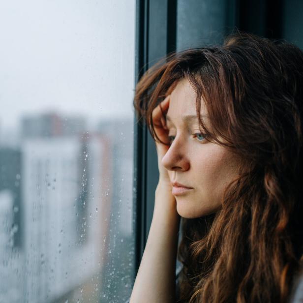 Nachdenkliche Frau mit langem Haar schaut regnerisch aus dem Fenster.