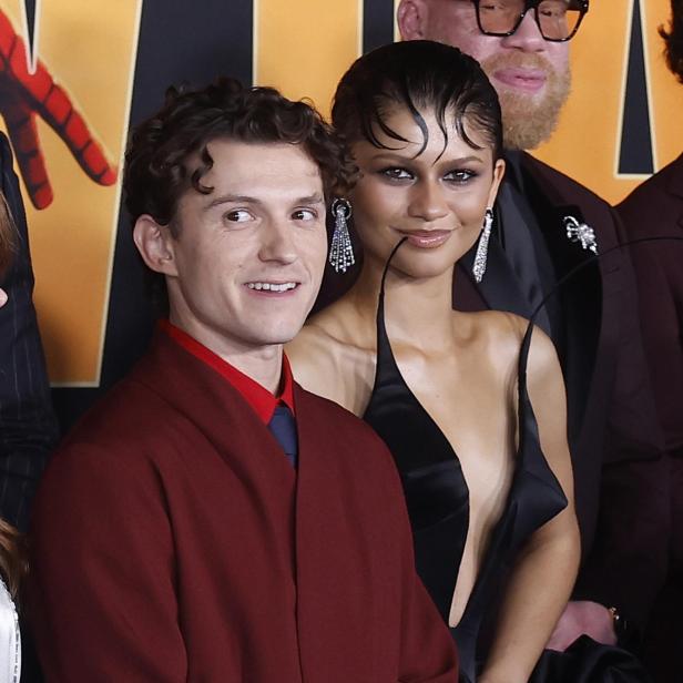 Tom Holland in einem weinroten Anzug und Zendaya in einem schwarzen Kleid posieren gemeinsam vor einem Hintergrund.