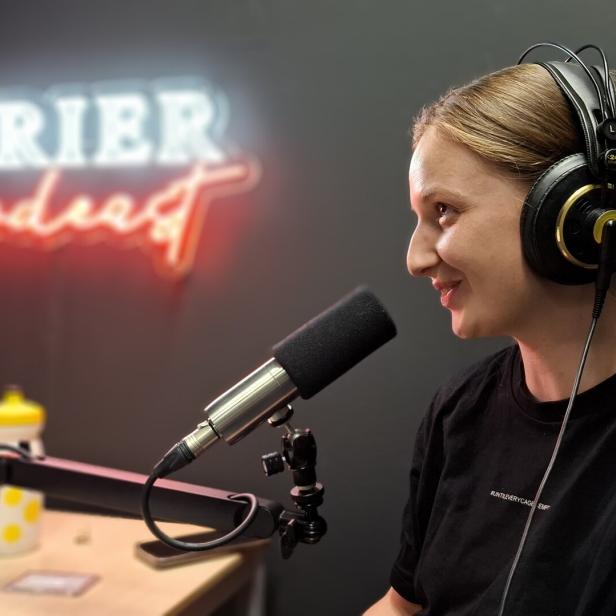 Eine Frau mit Kopfhörern spricht vor einem Mikrofon in einem Studio mit einem leuchtenden „KURIER podcast“-Schriftzug im Hintergrund.