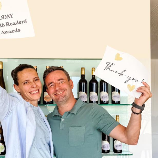 Ein lächelndes Paar hält Schilder mit der Aufschrift „USA TODAY 10Best's 2026 Readers' Choice Awards“ und „Thank you!“ vor einem Weinregal.