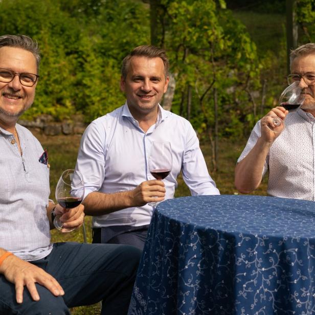 Drei Männer sitzen draußen an einem Tisch, trinken Rotwein und lächeln in die Kamera.