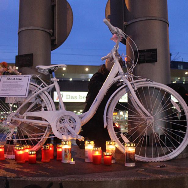 Weiße Gedenkfahrrad-Skulptur mit Kerzen am Fuß einer Brücke bei Dämmerung.