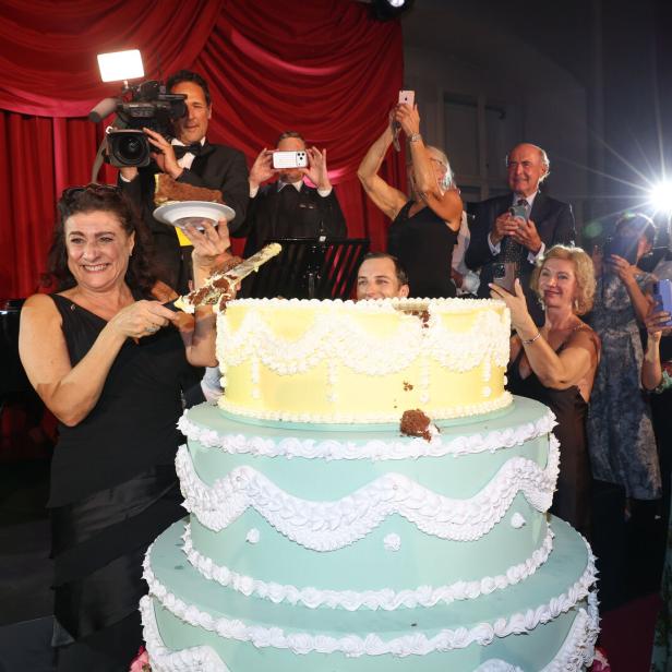 Eine Frau schneidet vor rotem Vorhang eine riesige Torte an, umgeben von applaudierenden Gästen und Fotografen.