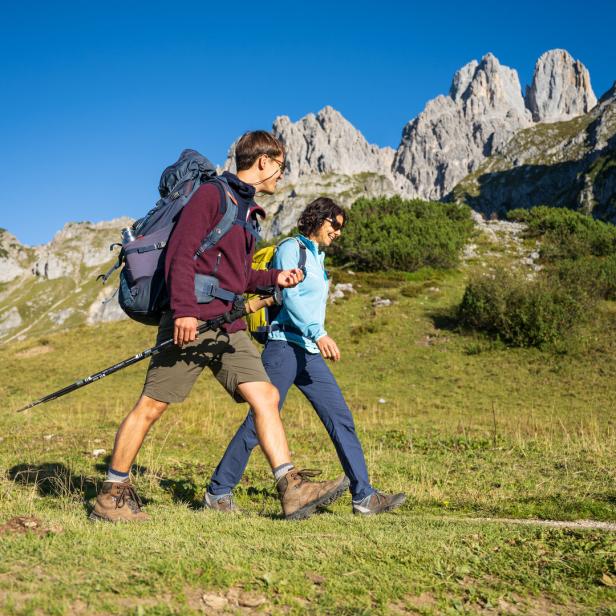 Zwei Wanderer mit Rucksäcken und Stöcken gehen über eine Wiese vor felsigen Bergen.