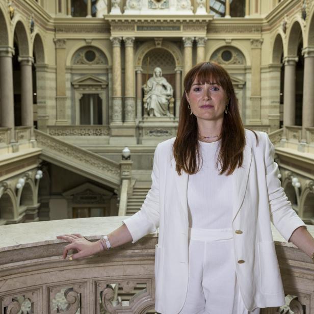 Frau in weißem Anzug steht in einer prunkvollen Halle mit Säulen und Treppe.