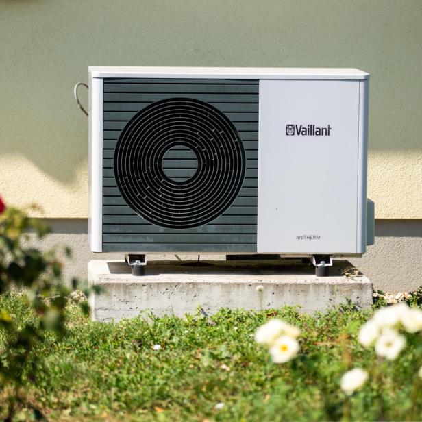 Vaillant-Wärmepumpe auf einem Betonsockel vor einer Hauswand, im Vordergrund unscharfe Blumen und Rasen.