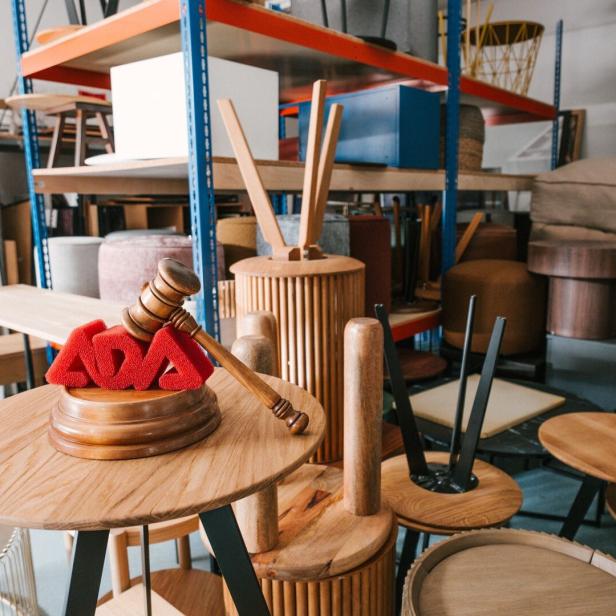 Ein Lager voller gestapelter Holzmöbel und Stühle, vorne ein Holzhammer auf einem Tisch mit roten ADA-Buchstaben.