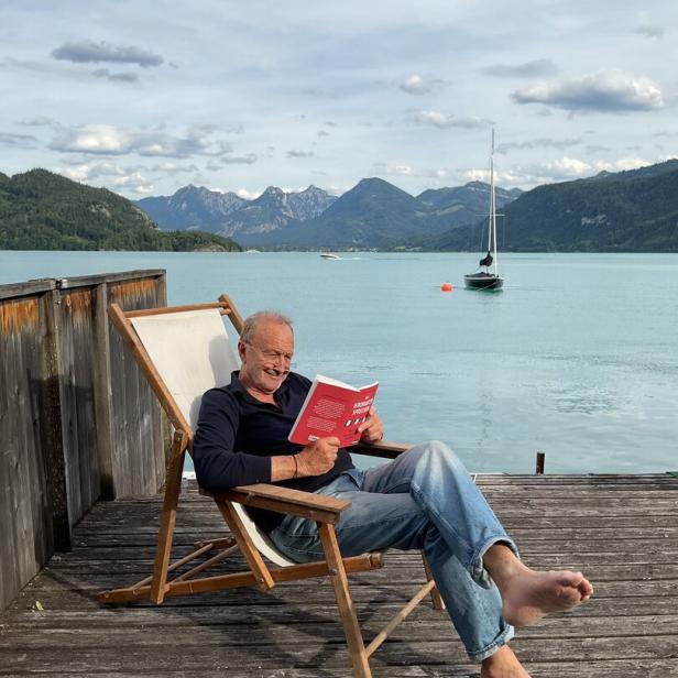 Ein Mann liest entspannt auf einer Holzterrasse am See, mit Segelboot und Bergen im Hintergrund.
