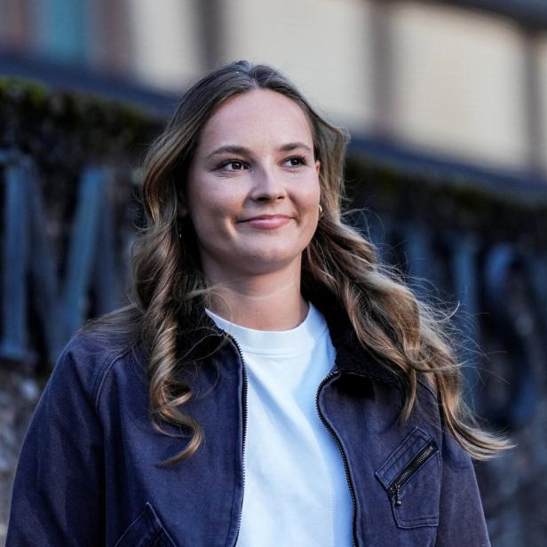Ingrid Alexandra mit langem, gewelltem Haar trägt eine dunkle Jacke über einem weißen Shirt und lächelt vor einer steinernen Wand.