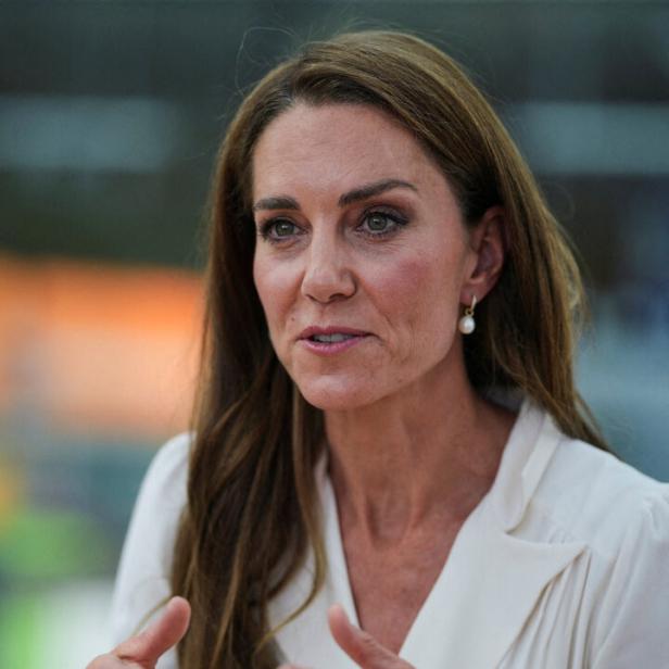 Prinzessin Kate spricht gestikulierend in einem weißen Blazer mit Perlenohrringen.