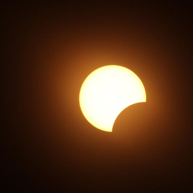 Teilweise verfinsterte Sonne mit dunkler Sichel vor orangefarbenem Himmel.