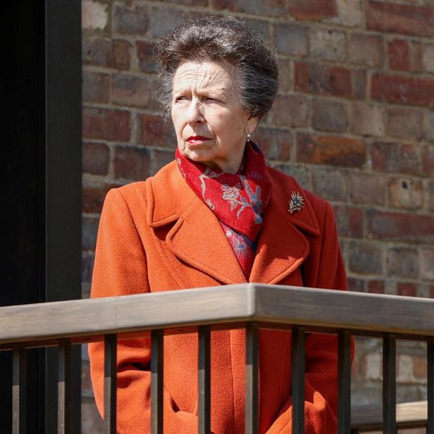Prinzessin Anne trägt einen orangefarbenen Mantel und ein gemustertes Tuch vor einer Backsteinmauer.