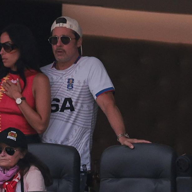 Brad Pitt trägt eine Sonnenbrille und ein USA-Trikot, während er neben einer Frau in einem roten Oberteil auf einer Tribüne steht.
