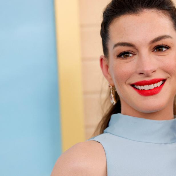 Anne Hathaway lächelt mit rotem Lippenstift und trägt ein hellblaues Oberteil vor einem blau-beigen Hintergrund.