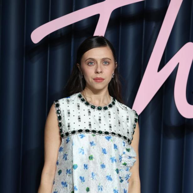 Bel Powley trägt ein weißes, ärmelloses Kleid mit blauem Blumenmuster und grünen Schmucksteinen vor einem dunklen Hintergrund.