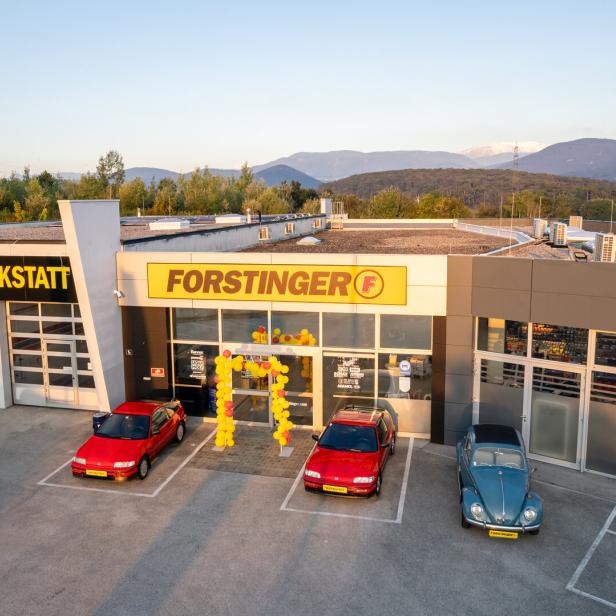 Vor einem Autohaus mit den Schildern „FACHWERKSTATT“ und „FORSTINGER“ stehen drei Autos, davor ein gelber Ballonbogen.