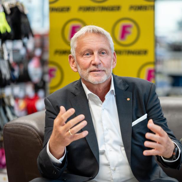 Älterer Mann im Anzug spricht gestikulierend auf einem Sofa in einem Laden, mit gelbem F‑Hintergrund.